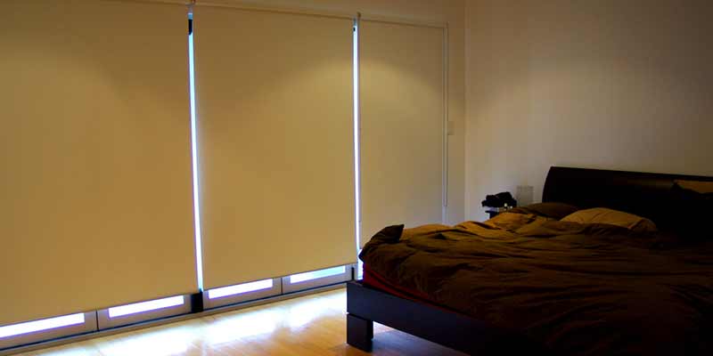 foto-secundaria-telas-blackout-002 Habitación moderna con una cama oscura, y ventanales cubiertos por persianas enrollables con tejidos blackout