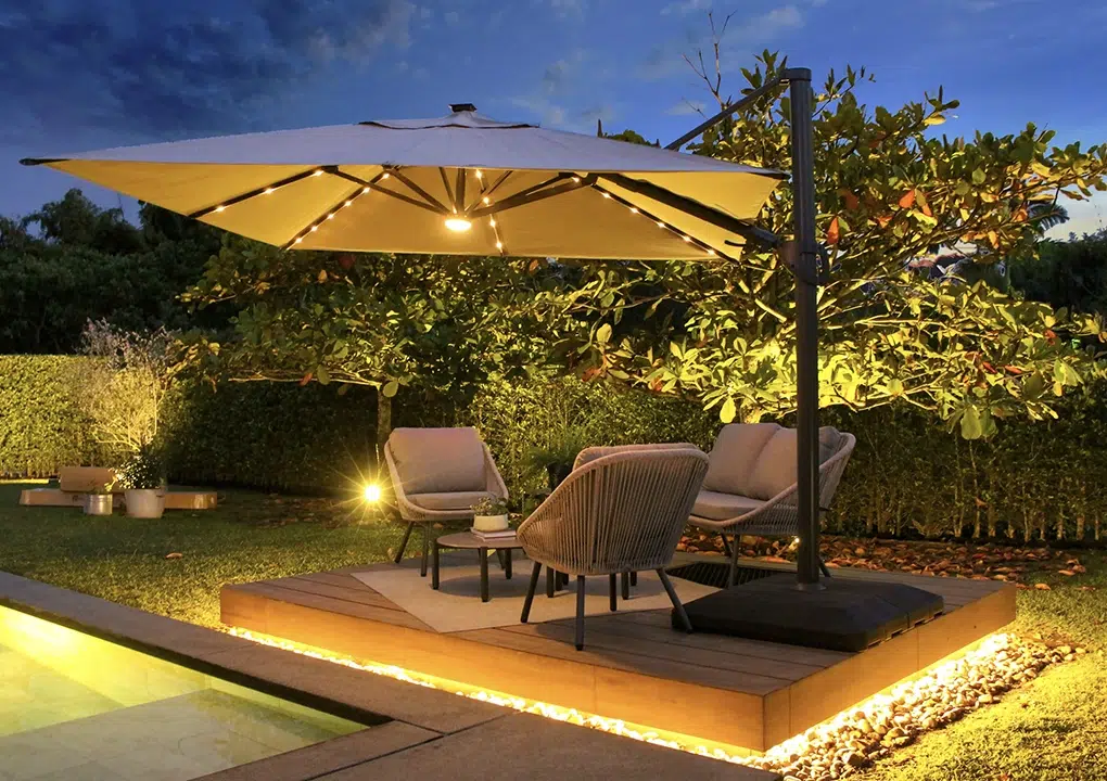 Sombrilla Marea en color beige abierta de noche con la luces LED prendidas en una casa campestre en el área de la piscina en una sala pequeña de exterior