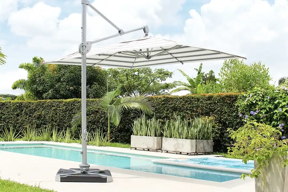 Sombrilla Cadiz blanca de mástil lateral junto a una piscina alargada, con seto recortado y maceteros de concreto