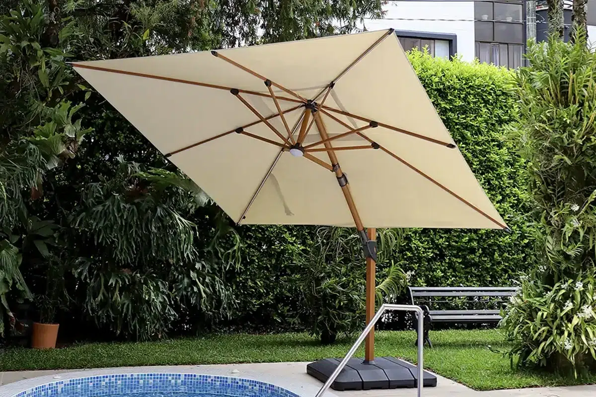 Sombrilla Kiosk Wood beige con mástil color madera junto a una piscina de mosaico azul, rodeada de jardín