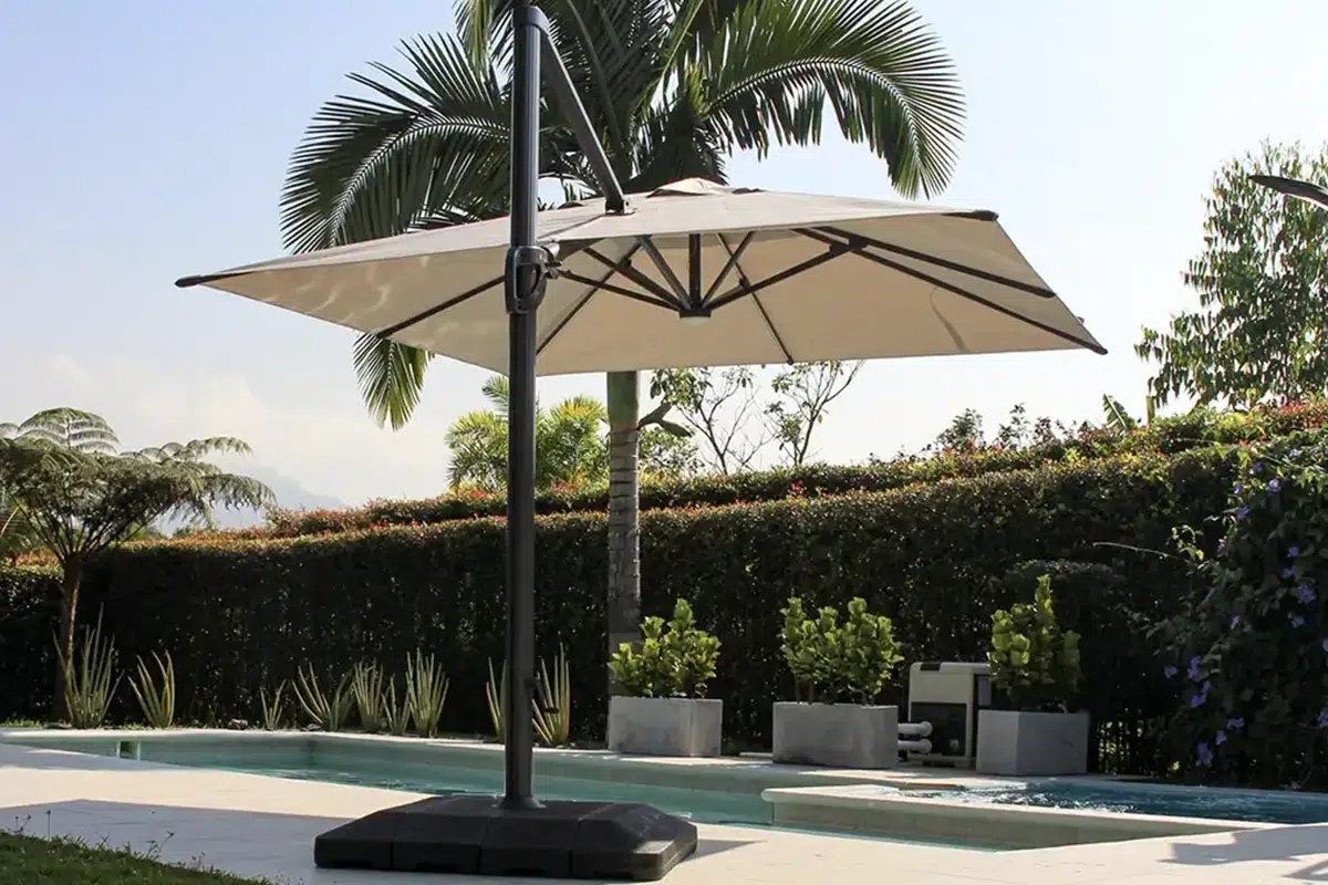 Sombrilla Marea beige con estructura antracita junto a una piscina, entre palmeras y maceteros de concreto