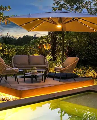Sombrilla Marea iluminada de noche sobre un deck con sofás, en un jardín con luz cálida