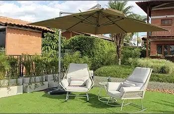 Sombrilla Cadiz beige sobre el césped con dos sillones, frente a una casa de ladrillo