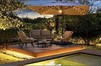 Sombrilla Marea encendida al anochecer sobre un deck con sofás, junto a la piscina